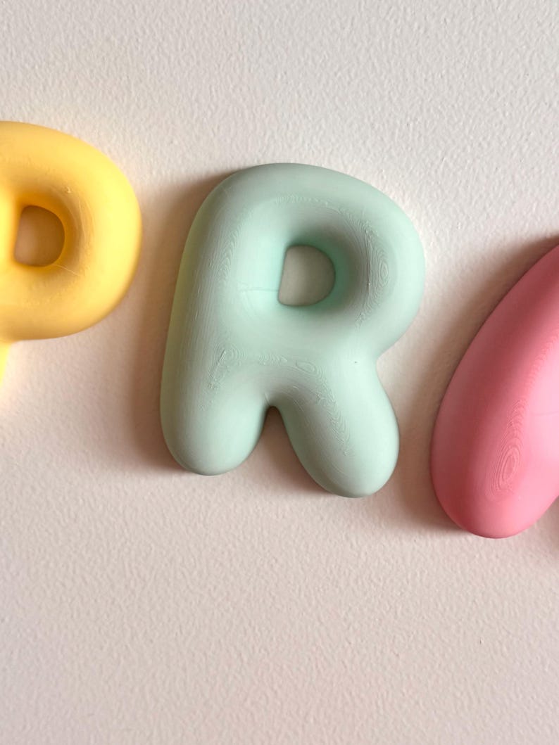 May include: A three-dimensional letter P in a soft mint green color with a rounded, bubble-like shape. The letter is positioned between a partial yellow letter to the left and a partial pink letter to the right, all featuring a smooth finish.