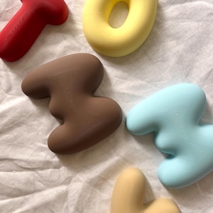 May include: Five smooth, rounded, three-dimensional objects in various colors including red, yellow, brown, light blue, and beige. Each piece features a soft, matte finish with subtle, concentric circular textures visible on the surface.