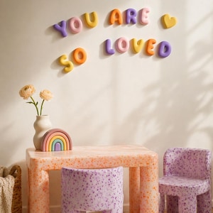 May include: A children's table and two chairs with a speckled orange and purple finish. On the wall, colorful 3D letters spell out YOU ARE SO LOVED with a heart icon. A small rainbow-shaped object and a vase with flowers sit on the table.