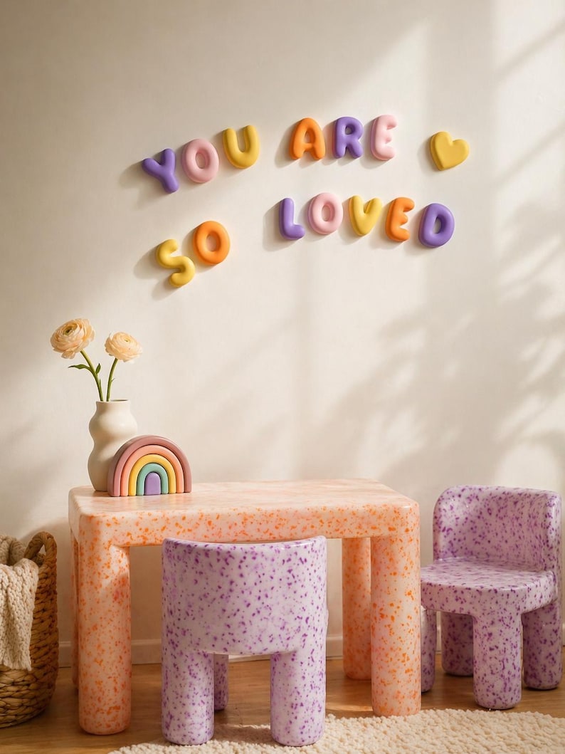 May include: A children's table and two chairs with a speckled orange and purple finish. On the wall, colorful 3D letters spell out YOU ARE SO LOVED with a heart icon. A small rainbow-shaped object and a vase with flowers sit on the table.