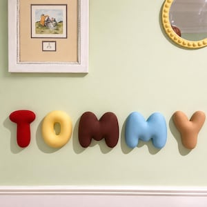 May include: A set of five plush, rounded, three-dimensional letters spelling the name TOMMY. The letters are coloured red, yellow, dark brown, light blue, and tan, and are mounted on a light green wall below a framed picture and a round mirror.