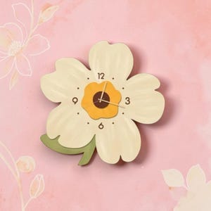 May include: A floral wall clock with a cream-coloured face and a yellow centre. The clock has black numbers at the 12, 3, 6, and 9 o'clock positions. A green leaf is attached to the bottom left of the clock. The background is pink.