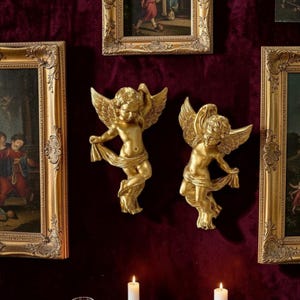 Op de afbeelding: Twee goudkleurige engelenbeelden voor aan de muur met uitgespreide vleugels en vloeiende stof. Ze zijn bevestigd op een dieprode fluwelen muur, omringd door ingelijste schilderijen en verlicht door kaarsen.