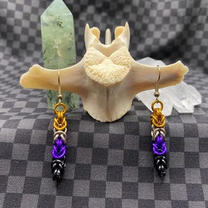 May include: A pair of handmade chainmail earrings with gold, silver, purple, and black rings. The earrings are displayed on a bone-shaped object against a checkered background. The earrings have a unique, geometric design.