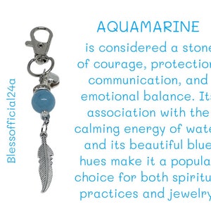 May include: A silver keychain with a blue aquamarine stone bead, a silver feather charm, and a silver ring. The text "AQUAMARINE" is written in blue letters above the text "is considered a stone of courage, protection, communication, and emotional balance. Its association with the calming energy of water and its beautiful blue hues make it a popular choice for both spiritual practices and jewelry."