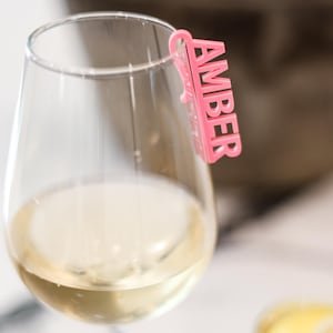 May include: A glass of white wine with a pink plastic glass marker that says "Amber".