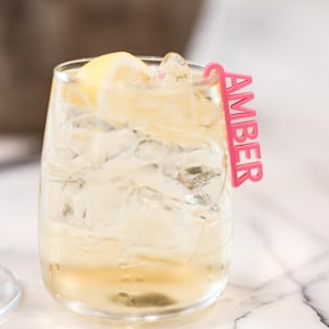 May include: A clear glass filled with a light-colored beverage, ice cubes, and a lemon wedge. A bright pink acrylic drink stirrer with the name AMBER in capital letters is hooked onto the rim of the glass, extending vertically upward.