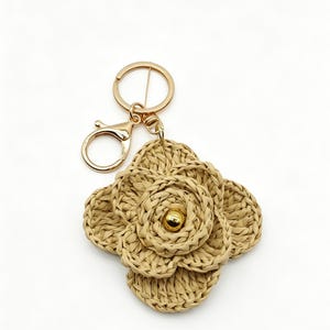 May include: A beige crocheted flower keychain featuring a layered petal design with a central gold-tone metallic bead. The floral charm is attached to a gold-tone metal split ring and a lobster claw clasp, providing a secure way to attach keys.