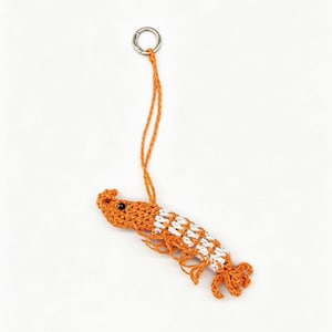 May include: A crocheted shrimp keychain featuring orange and white yarn, a small black bead for an eye, and a silver-toned metal ring attached to a long orange yarn loop for hanging. The shrimp has detailed legs and a segmented tail structure.