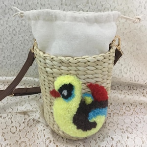 May include: A woven straw bucket bag with a white drawstring closure and a brown shoulder strap. The bag features a yellow, blue, red, and brown duckling applique on the front. The bag is designed to be a crossbody bag.