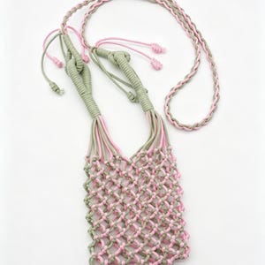 May include: A small crossbody bag crafted from sage green and light pink cord in a macrame net pattern. The bag features a long, braided shoulder strap made of the same two-tone cord, finished with knotted ends and adjustable sliding knots.