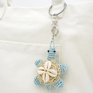 Puede incluir: Un llavero artesanal con diseño de tortuga. La tortuga tiene un cuerpo azul claro, un caparazón beige adornado con conchas marinas y está sujeta a un cierre metálico plateado. El llavero cuelga de un bolso tote blanco.