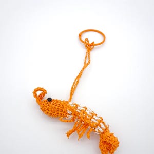 May include: An orange and white crocheted shrimp keyring with a black eye. The shrimp has an orange loop at the top for attaching to keys or a bag. The shrimp is approximately 10 cm long.