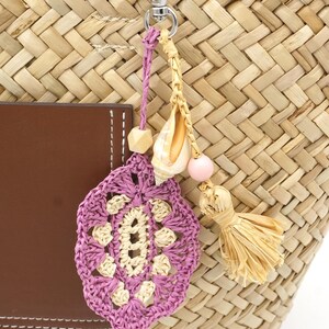 Pode incluir: Uma bolsa de palha trançada com um remendo de couro castanho e um pendente decorativo. O pendente inclui um motivo de croché roxo e creme, uma concha natural, uma conta rosa claro, uma conta de madeira e uma pequena borla de palha.