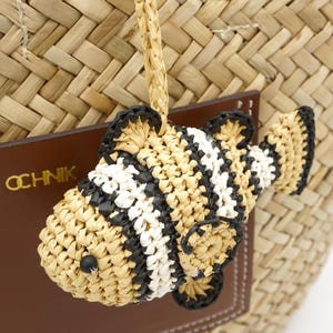 May include: A woven straw fish-shaped charm with black and white stripes, attached to a woven straw bag. The fish has black accents and a small black bead for an eye. A brown leather tag with the word "OCHNIK" is visible.