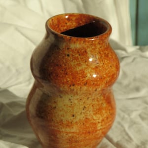 Puede incluir: Un jarrón de cerámica con silueta de doble bulbo y acabado brillante. La superficie presenta un esmalte moteado en tonos cálidos naranja, óxido y crema, acentuado por pequeñas motas de color marrón oscuro en todo el exterior del recipiente.