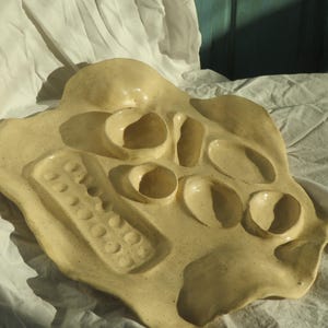 May include: A cream-coloured ceramic serving tray with a speckled finish and an irregular, organic shape. The tray features multiple recessed compartments of varying sizes and shapes, including a long rectangular section with small circular indentations.