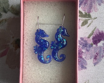 May include: A pair of blue seahorse earrings with iridescent glitter accents, displayed in a pink box with white foam padding. The earrings have silver-colored hooks.