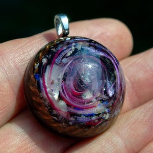 May include: A circular resin pendant featuring a swirling design of deep purple, magenta, and white pigments embedded with metallic copper wire coils and small crystalline fragments, finished with a silver-toned metal bail at the top.
