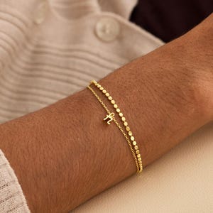 May include: A delicate gold bracelet with two fine chains. One chain features small, round beads, while the other has a small letter "H" charm. The bracelet is worn on a person's arm, against a neutral background.
