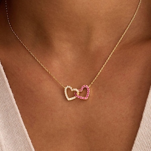 May include: A gold necklace with two intertwined heart pendants. One heart is set with clear crystals, the other with pink crystals. The necklace is worn on a person's chest, highlighting the fine chain and pendant design.