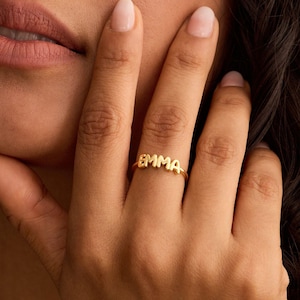 May include: A gold ring spelling "EMMA" in capital letters. The ring is on a finger, with a hand and part of a person's face visible. The ring is a delicate piece of jewellery. The image is a close-up shot of the ring.