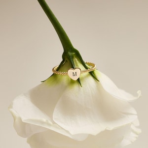 Peut inclure: Une bague dorée avec un charm en forme de cœur portant la lettre "M" repose sur une fleur blanche. La bague a un design de bande torsadée. La tige de la fleur est verte et les pétales sont blancs. L'arrière-plan est de couleur neutre.