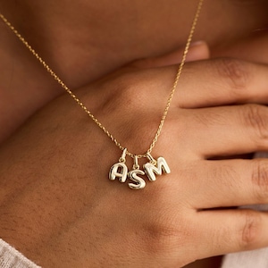 May include: A gold-colored necklace with three initial charms spelling out "ASM". The necklace is delicate, with a thin chain and rounded letter pendants. The letters are in a serif font and hang close together.