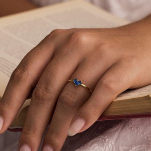 Könnte beinhalten: Ein goldener Ring mit herzförmigem Design, der zwei blaue Edelsteine ​​aufweist. Der Ring befindet sich am Finger einer Person, die auf einem offenen Buch ruht. Das zarte Band des Rings und die leuchtend blauen Steine ​​erzeugen einen auffälligen Kontrast.