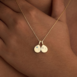 May include: A gold necklace featuring two teardrop pendants. Each pendant has a small star design and a single initial. One pendant has a blue star, and the other has a star-shaped design. The necklace is shown on a person.