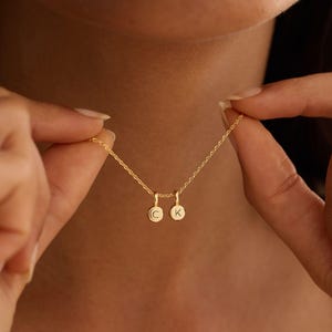 May include: A gold-toned necklace featuring a delicate chain and two small, circular charms. Each charm is engraved with a single letter, specifically the letters C and K, suspended from the chain in a side-by-side arrangement.