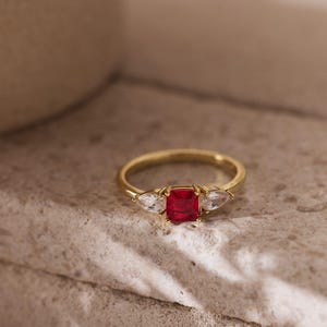 May include: A gold ring with a rectangular red gemstone center and two pear-shaped white gemstones on either side. The ring is set on a textured, light-colored surface.