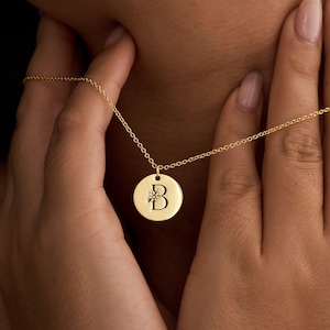 Puede incluir: Un collar dorado con un colgante circular. El colgante presenta la letra "B" con un diseño floral. El collar lo sostiene una persona. La cadena es delicada y el colgante está centrado.