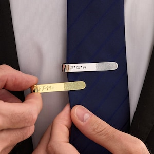 May include: Two rectangular tie clips, one silver-toned and one gold-toned, attached to a dark blue tie. The silver clip features the text 12 08 24 and a fingerprint. The gold clip features the text To The Moon and a fingerprint design.