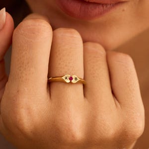 May include: A gold ring featuring two small heart-shaped segments joined together. The left heart is engraved with the letter A, and the right heart is engraved with the letter B. A small, round, vibrant red gemstone is set between the two hearts.
