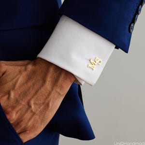 May include: A pair of gold-toned metal cufflinks featuring the letters M and S in a serif font. The cufflinks are attached to the white fabric cuff of a long-sleeved shirt, which is worn underneath a dark blue blazer with visible buttons on the sleeve.