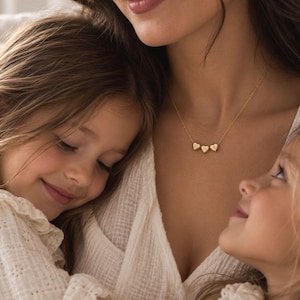 Puede incluir: Un collar de tono dorado con tres pequeños dijes en forma de corazón, cada uno grabado con una letra individual: E, H y K. Los dijes cuelgan de una delicada cadena de tono dorado que descansa sobre el cuello, creando un aspecto sencillo.