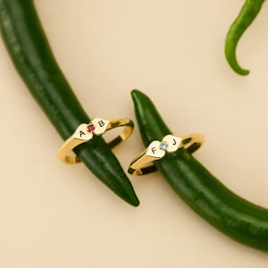 May include: Two gold rings featuring heart-shaped settings. One ring is engraved with the letters A and B and set with a small red stone. The other ring is engraved with the letters F and J and set with a small light blue stone.