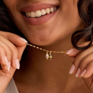 May include: A delicate gold necklace with two small gemstone pendants. One pendant is a clear, oval-shaped stone, and the other is a green, oval-shaped stone. The necklace is being held up by a person.
