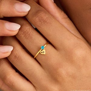May include: A thin gold ring featuring a small, round, bright blue gemstone and a tiny, dangling gold heart charm engraved with the letter M. The ring is constructed from a slender metallic band that encircles the finger, showcasing a minimalist design.