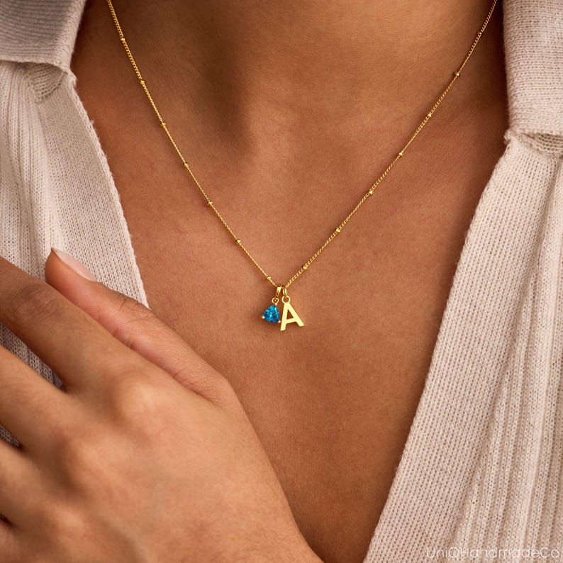 May include: A gold-colored necklace with a pendant featuring the letter "A" and a small blue gemstone. The necklace is delicate, with small beaded accents along the chain. The pendant hangs near the center of the chest.