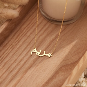 May include: A gold necklace with a delicate chain and a pendant featuring the Arabic word "Salam" in a simple, elegant font.