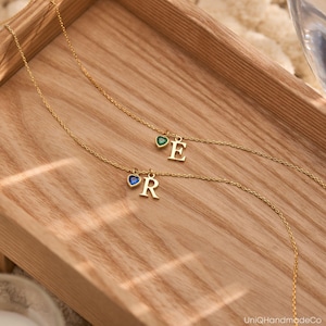 May include: Two gold necklaces with initial charms. The necklace on the left has a blue heart with the letter "R" and the necklace on the right has a green heart with the letter "E".