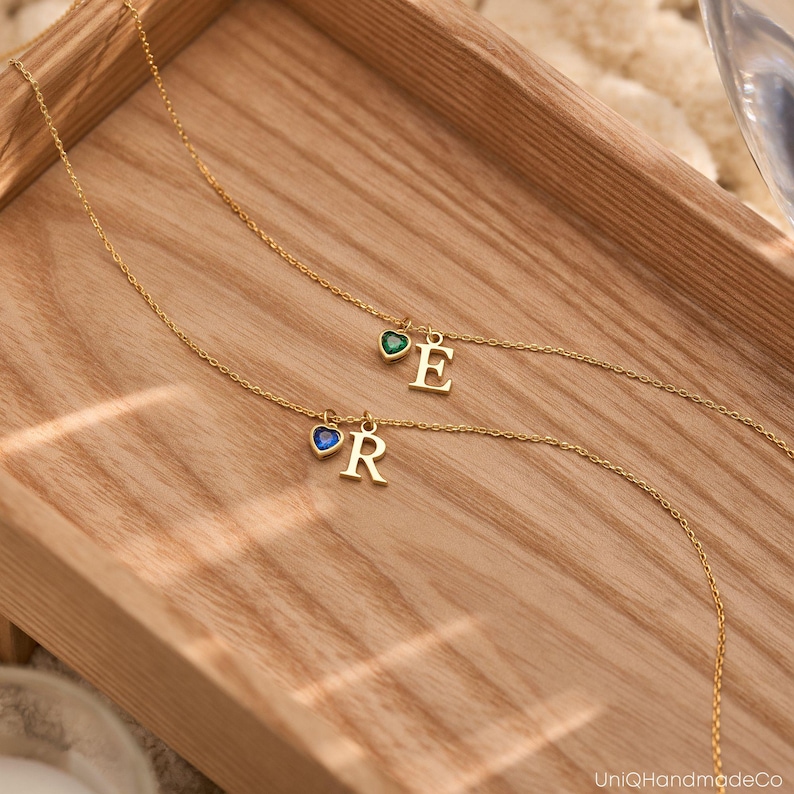 May include: Two gold necklaces with initial charms. The necklace on the left has a blue heart with the letter "R" and the necklace on the right has a green heart with the letter "E".