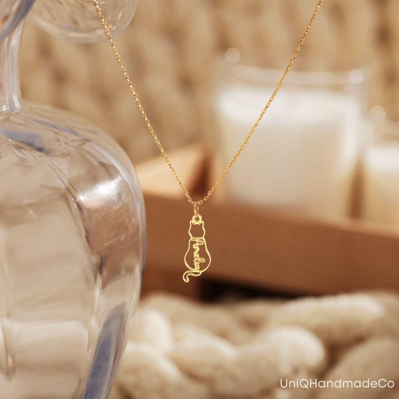 May include: Gold necklace featuring a cat-shaped pendant. The pendant is cut out and the word "lovely" is inscribed within the cat's body.