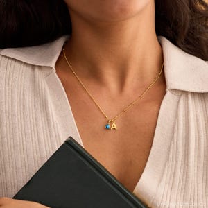 May include: A gold-colored necklace with a small blue gemstone and the letter "A" pendant. The necklace is worn on a person wearing a light beige shirt. The necklace is a delicate piece of jewelry.
