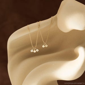 May include: Two gold-toned necklaces featuring small circular charms. One necklace has three charms engraved with the letters E, I, and B. The second necklace has two charms engraved with the letters C and K. The chains are delicate and gold-colored.