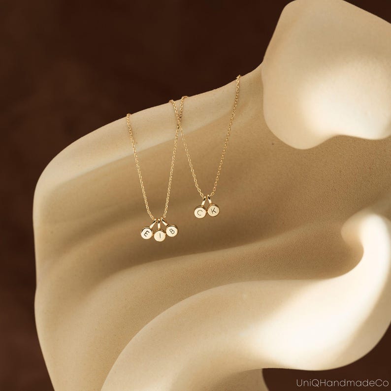 May include: Two gold-toned necklaces featuring small circular charms. One necklace has three charms engraved with the letters E, I, and B. The second necklace has two charms engraved with the letters C and K. The chains are delicate and gold-colored.