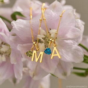 May include: Three gold necklaces with pendant letters "M" and "A". Each necklace has a small, colorful gemstone pendant. The necklaces are displayed against a backdrop of soft pink and white flowers. The image is well-lit and focuses on the jewelry.