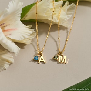 May include: Two gold-colored necklaces with delicate chains and small, round beads. Each necklace features a pendant with a letter and a gemstone. One has a blue gemstone and the letter "A", the other has a green gemstone and the letter "M".